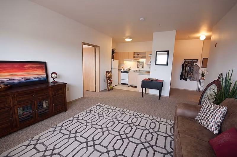 Interior view of a senior living apartment showing a living room area with a brown couch and patterned rug, a wooden cabinet with a framed sunset picture and a clock, and an open kitchen with white appliances and light wood cabinets. There is a small table and a coat rack with hanging clothes near the entrance.