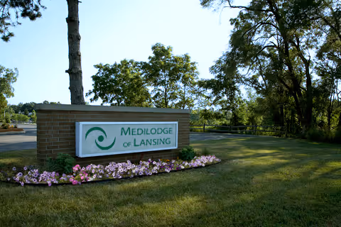 A brick sign for Medilodge of Lansing surrounded by a flower bed with pink flowers, set on a grassy area with trees and a clear sky in the background.