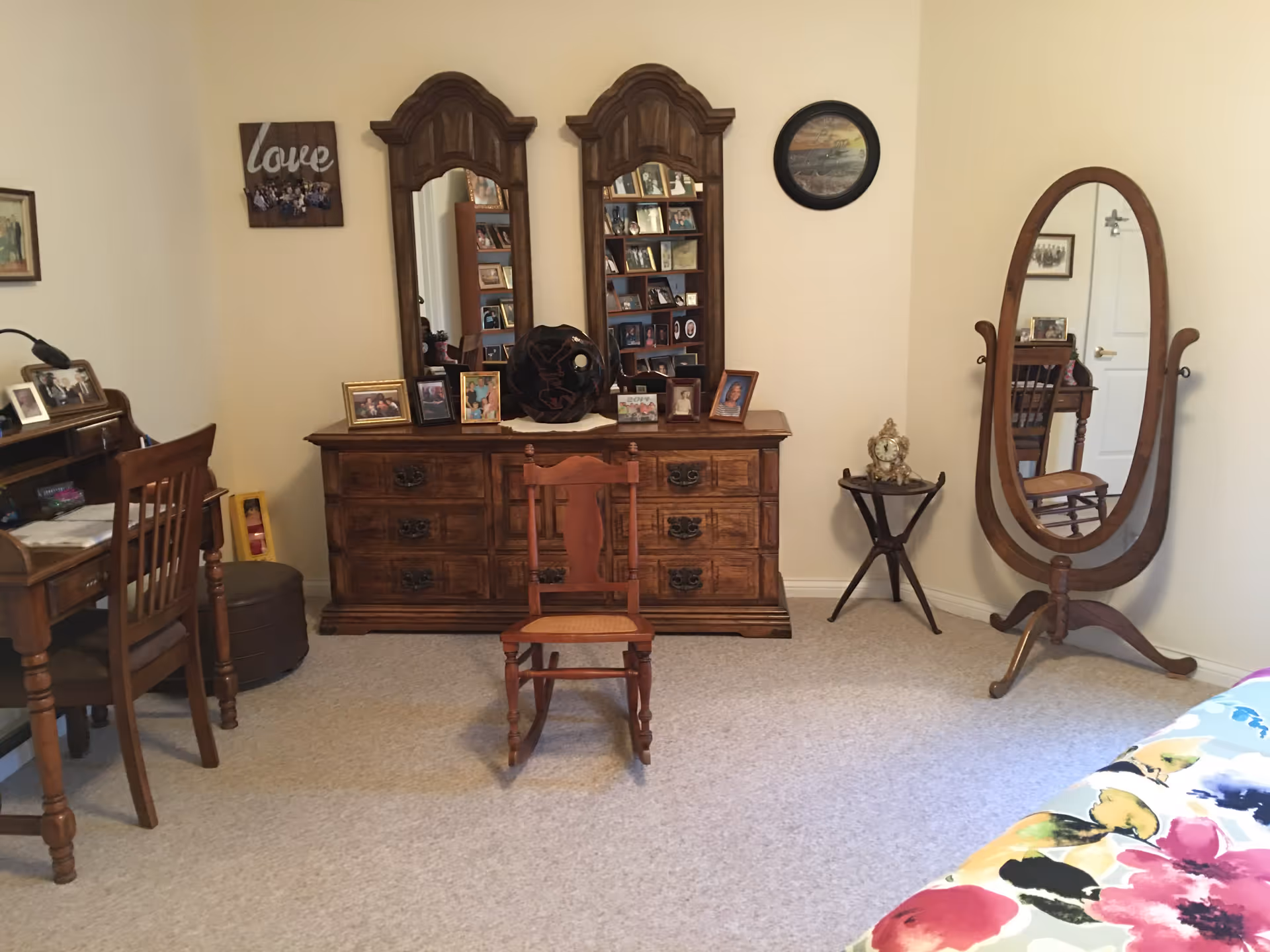 A cozy bedroom with a wooden dresser featuring two large mirrors and several framed photos on top. In front of the dresser is a wooden rocking chair. To the right, there is a wooden standing oval mirror and a small round table with a decorative clock. On the left side, there is a wooden desk with a chair and more framed photos. The bed with a colorful floral bedspread is partially visible on the right edge of the image. The walls are light-colored and decorated with a few pictures.