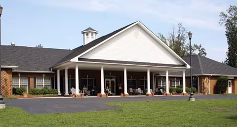 Front exterior view of a single-story brick building with a large covered porch supported by white columns, surrounded by green lawn and trees under a clear sky.