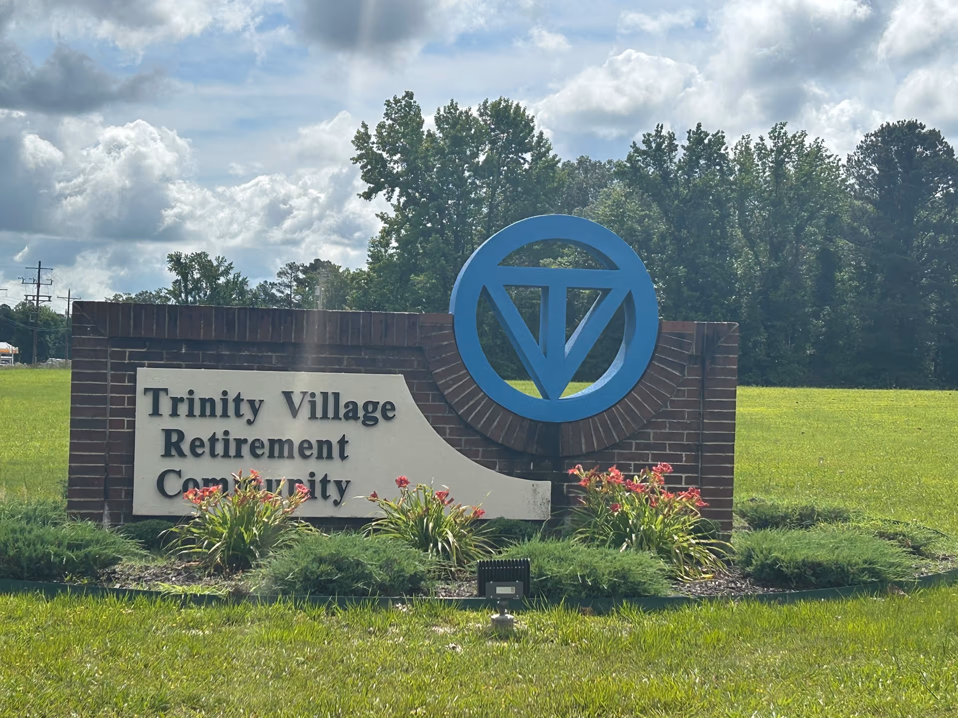 A brick sign for Trinity Village Retirement Community with a blue circular logo featuring a stylized 'TV'. The sign is surrounded by green grass, plants, and flowers, with trees and a partly cloudy sky in the background.