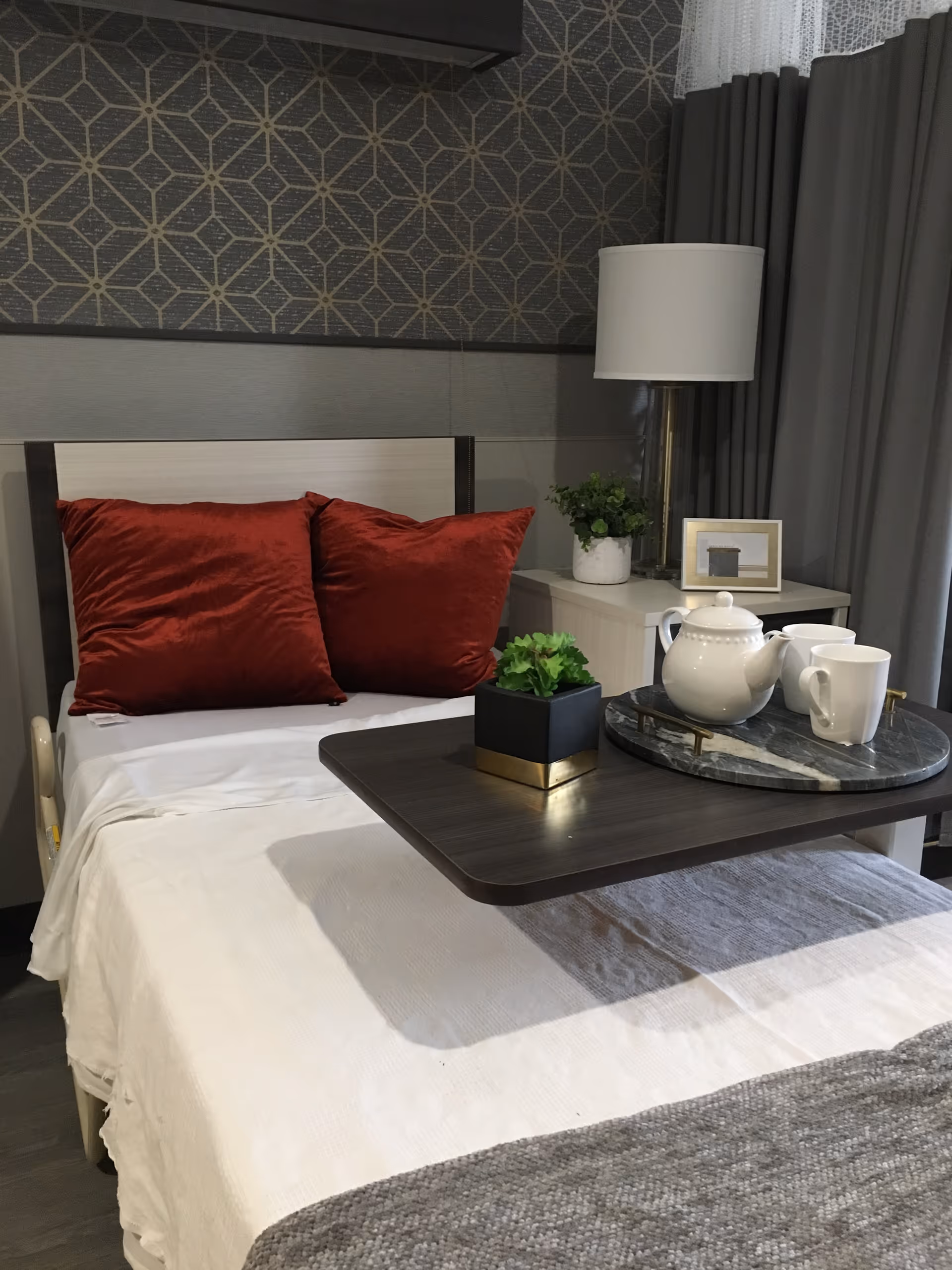 A neatly made single bed with white sheets and two red pillows against a patterned wall. A dark wooden overbed table holds a small potted plant and a round tray with a white teapot and two white mugs. Next to the bed is a nightstand with a lamp, a small plant, and a framed picture. Grey curtains and a lace curtain are visible in the background.