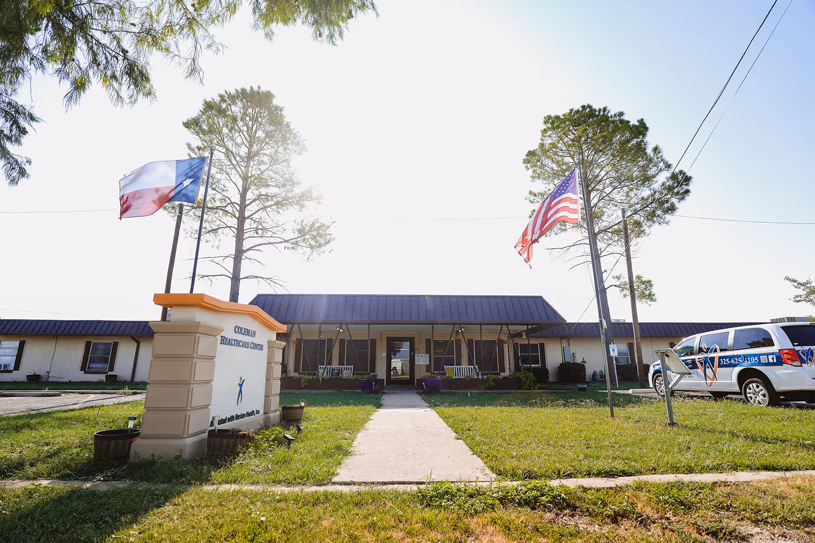 Front exterior view of Coleman Healthcare Center building with a sign in front, two flagpoles displaying the Texas and American flags, a sidewalk leading to the entrance, and a parked vehicle on the right side.