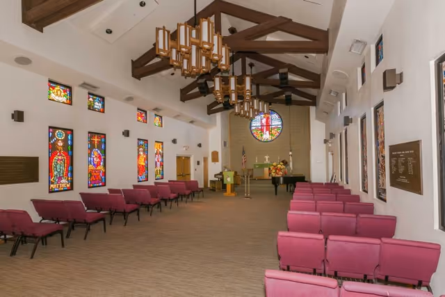 Interior of a chapel-like assembly room with rows of pink upholstered chairs, stained-glass windows, exposed wooden ceiling beams and an altar at the far end.