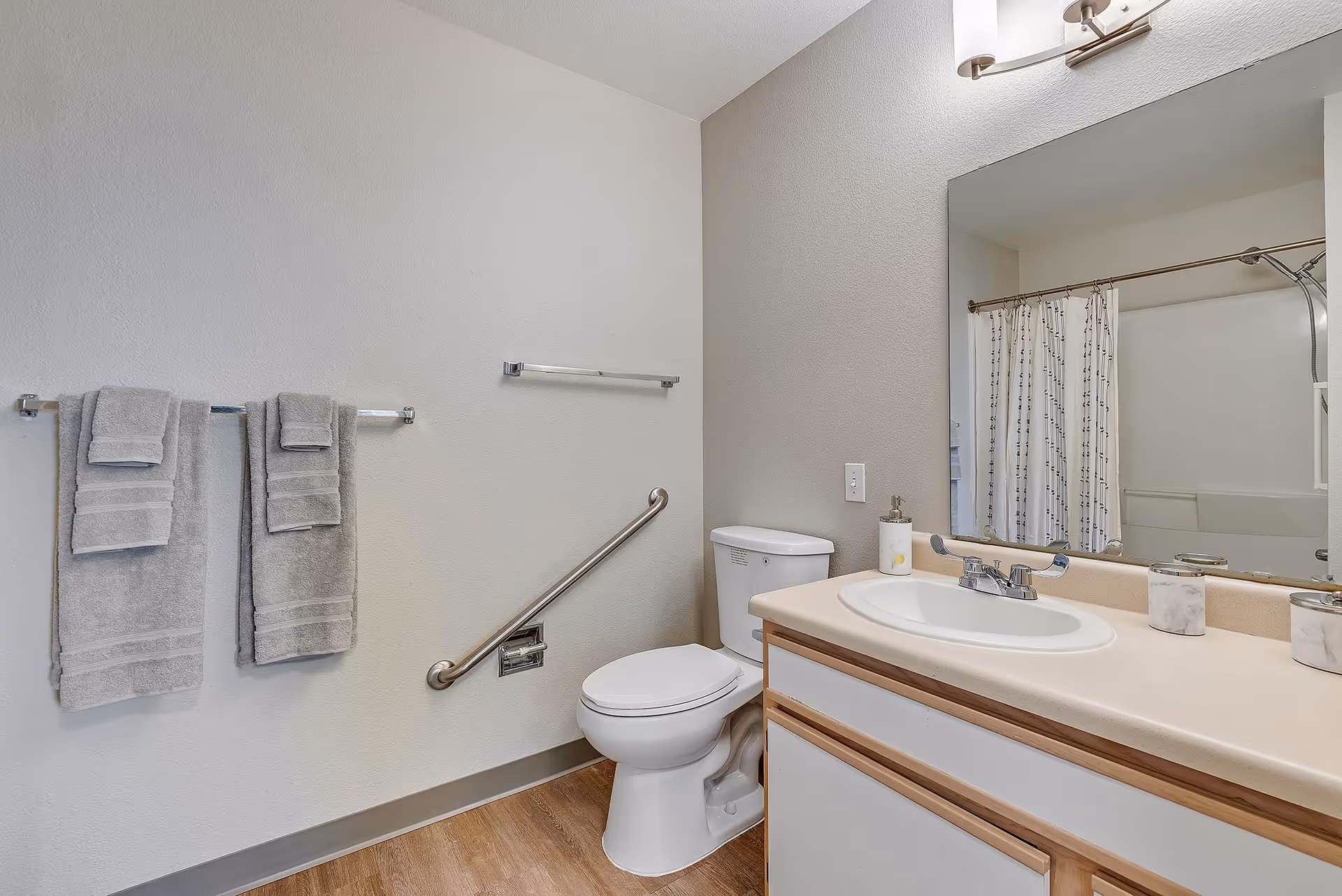 A clean and simple bathroom with a white toilet, a sink with a countertop, a large mirror above the sink, a shower with a curtain, and two gray towels hanging on a towel rack. There is a grab bar next to the toilet and wooden flooring.