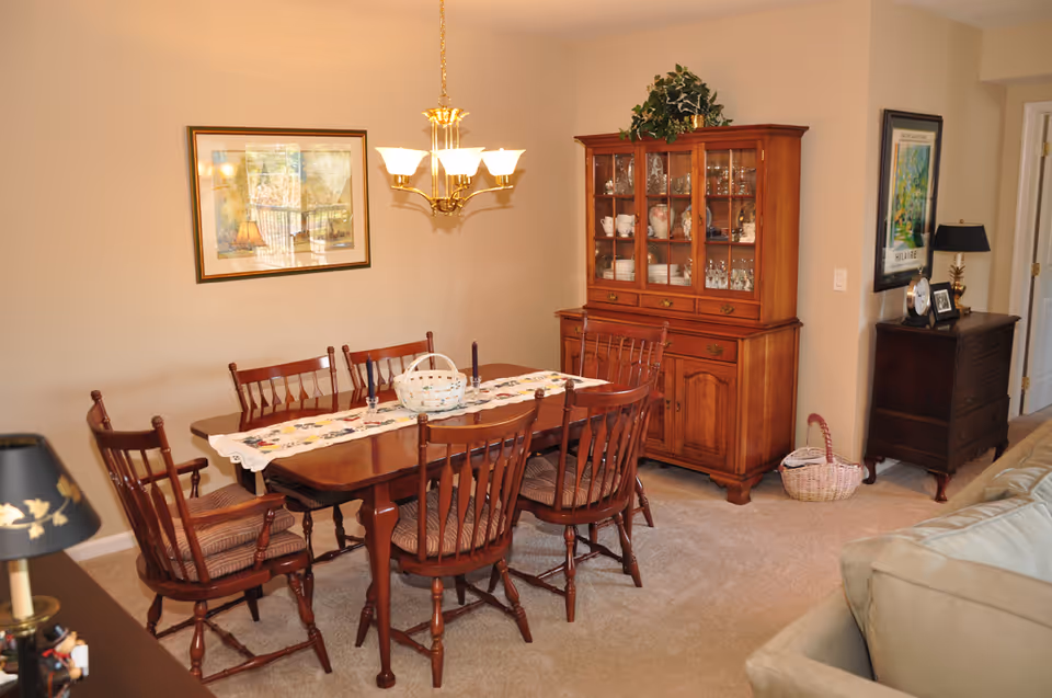A dining room with a wooden dining table surrounded by six matching wooden chairs with cushions. A decorative table runner and a white basket are on the table. A wooden china cabinet filled with dishes and glassware is against the wall. The room has beige walls, a framed picture, a chandelier light fixture, and carpeted floor. There is also a small dark wooden dresser with a lamp and framed photos on it, and a beige sofa partially visible in the foreground.