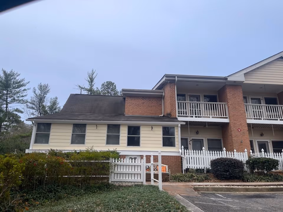 Two-story brick and beige-sided senior living building with balconies, a white picket fence, shrubs, and an adjacent parking area.