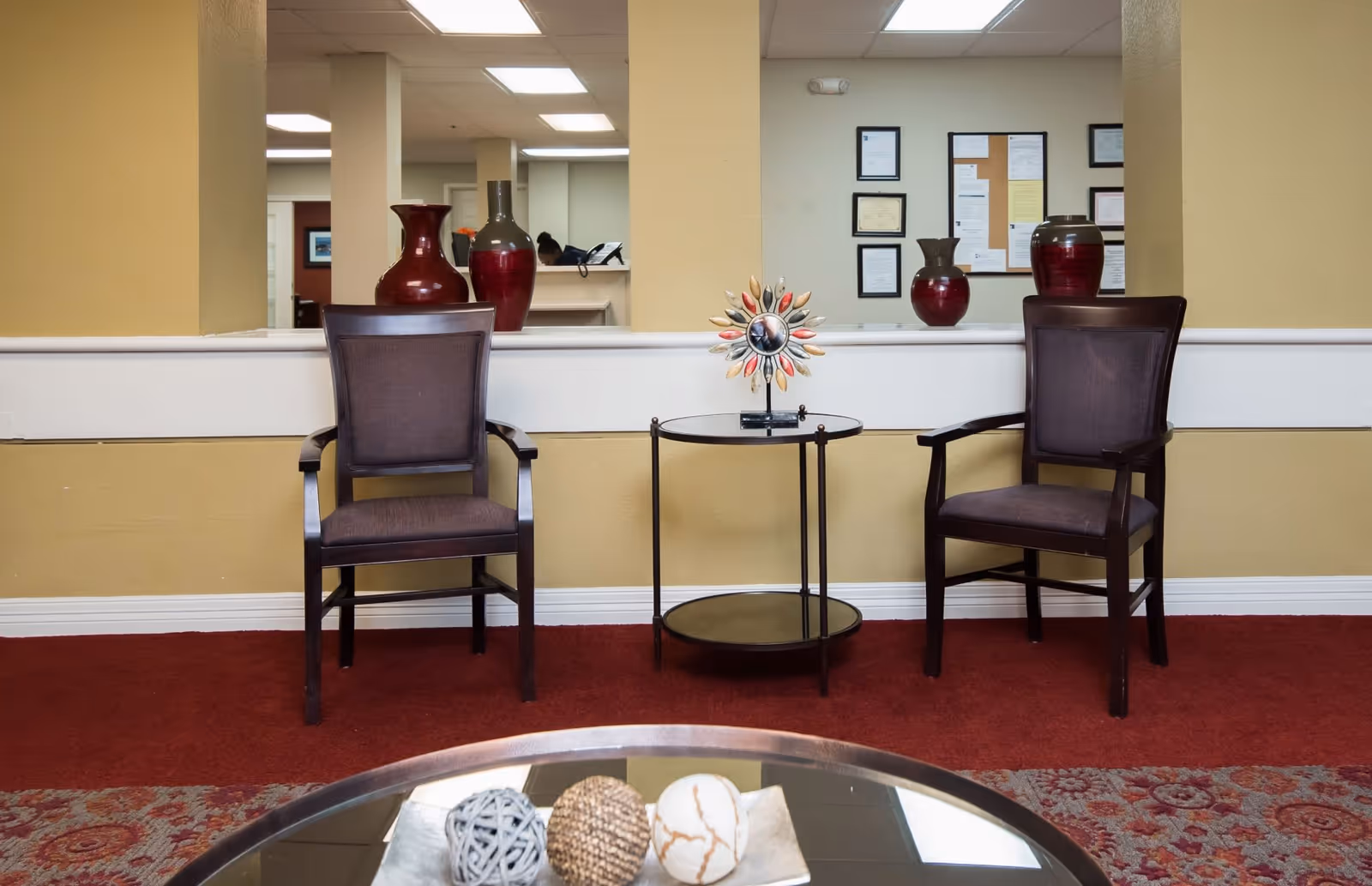Two wooden armchairs with dark cushions are placed on either side of a small round black table with a decorative sun-shaped ornament on top. Behind the chairs is a half wall with a large mirror reflecting framed certificates and a bulletin board on the opposite wall. The room has beige walls and red carpet flooring.