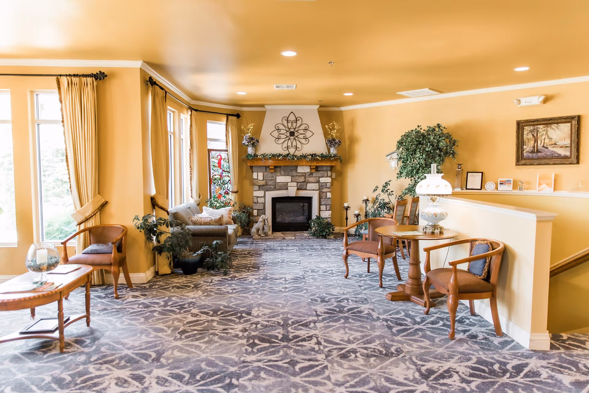 A cozy living room area with yellow walls and ceiling, patterned carpet, and a stone fireplace decorated with plants and a metal wall art piece above it. The room features several chairs, a small table with a decorative lamp, a sofa with cushions, large windows with curtains, and various potted plants. A framed painting hangs on the wall near a staircase.