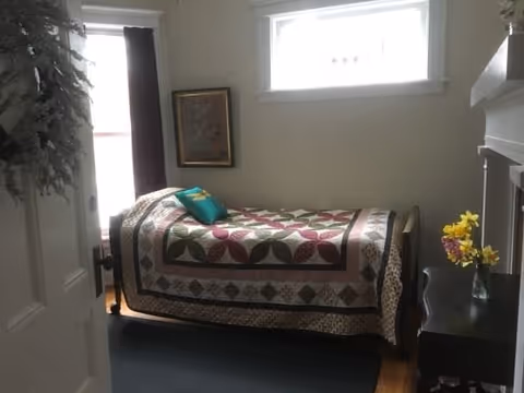 A small bedroom with a single bed covered by a colorful quilt featuring a geometric pattern. There is a teal pillow on the bed. A window with white trim is above the bed, letting in natural light. A framed picture hangs on the wall next to the window. A dark wooden nightstand with a vase of yellow flowers is beside the bed. The room has light-colored walls and a dark rug on the wooden floor.