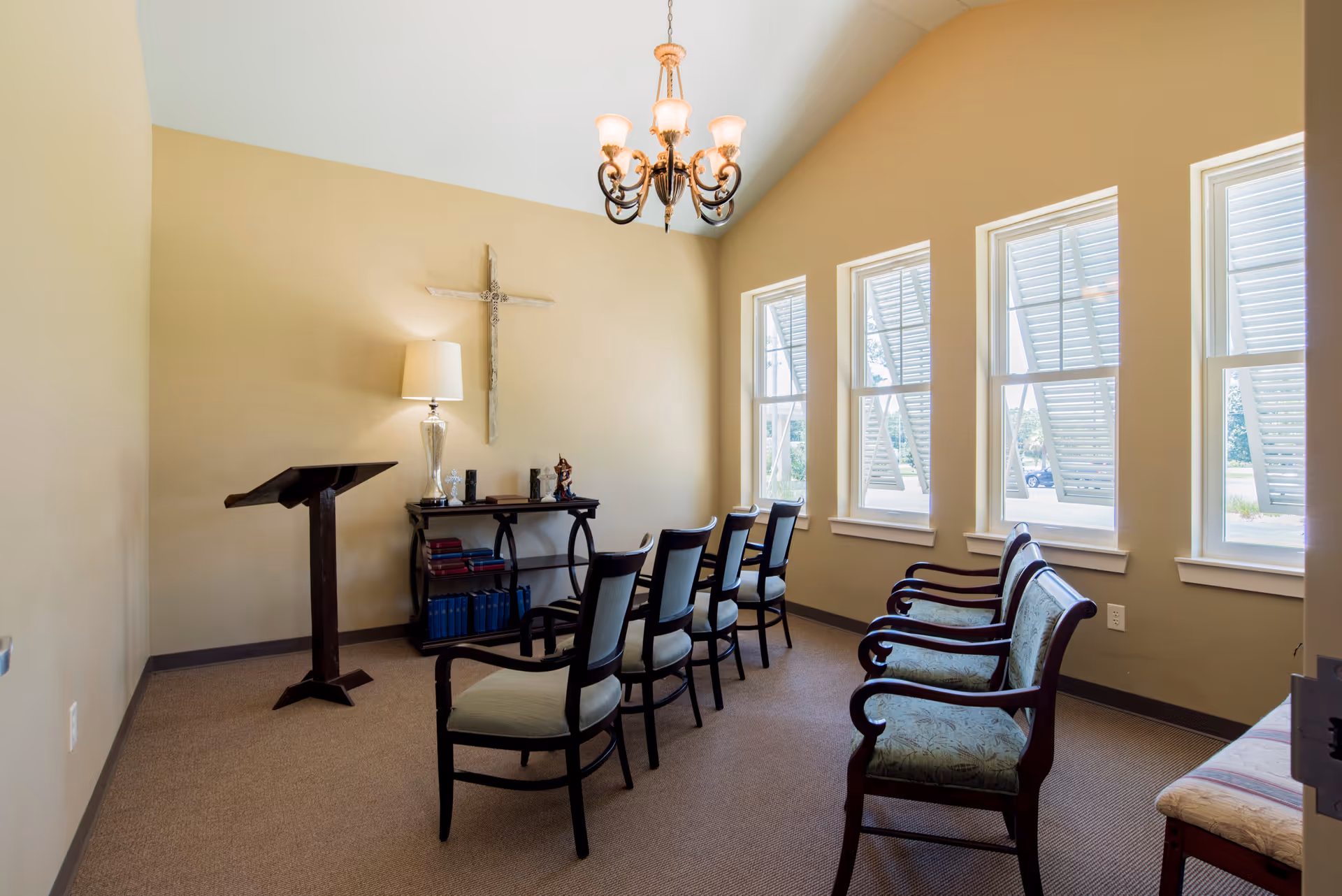 Small sunlit interior room with rows of chairs facing a lectern and a cross on the wall.