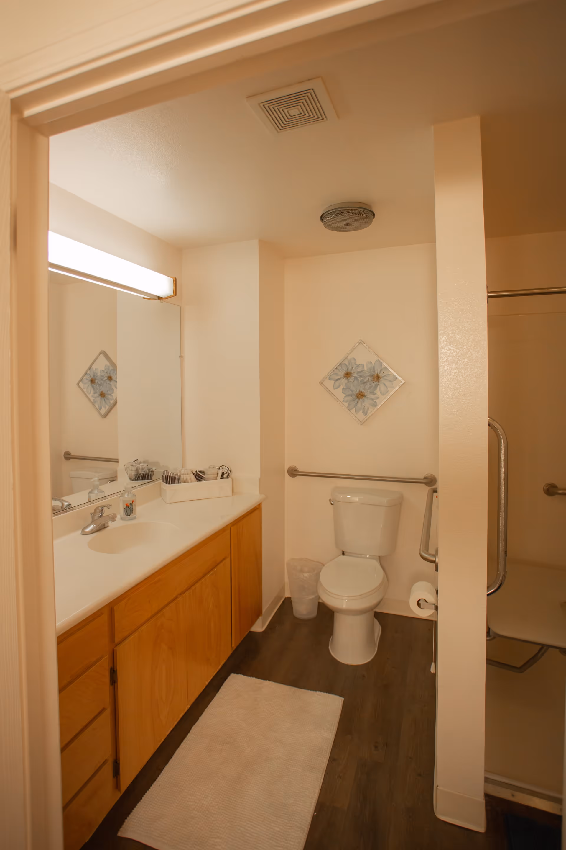 A clean bathroom with a wooden vanity and white countertop featuring a sink and a large mirror above it. There is a toilet next to the vanity with a grab bar on the wall behind it and a small trash can beside it. A white bath mat is placed on the wooden floor. On the right side, there is a shower area with a grab bar and a foldable shower seat. A decorative floral wall art hangs above the toilet.