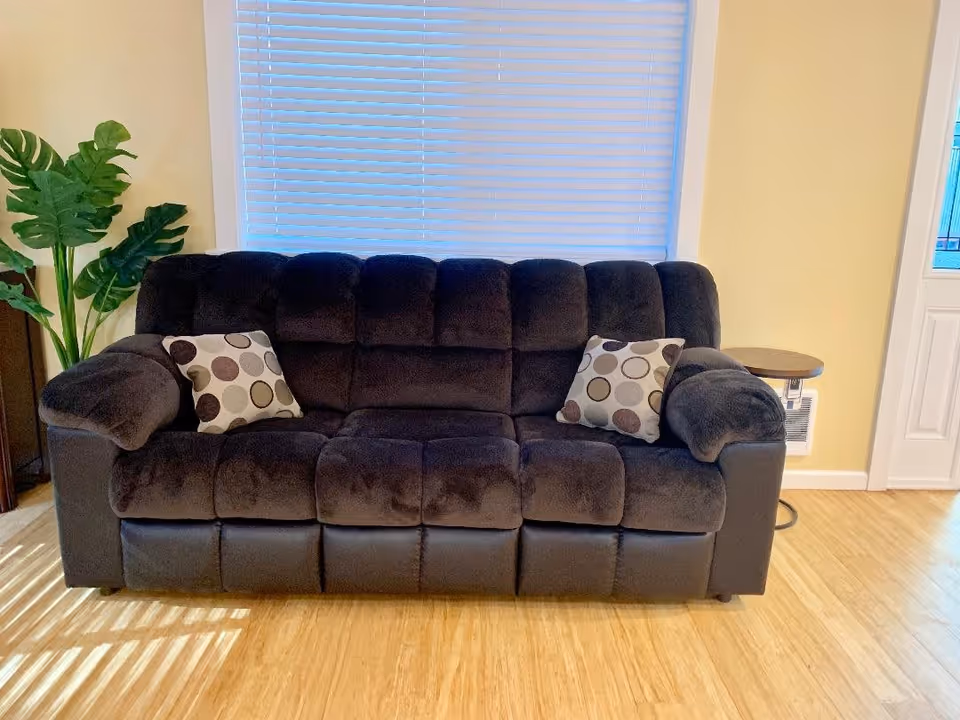 A dark brown plush sofa with two patterned throw pillows placed in front of a window with closed blinds. To the left of the sofa is a green leafy plant, and to the right is a small round wooden side table. The room has light yellow walls and a wooden floor.
