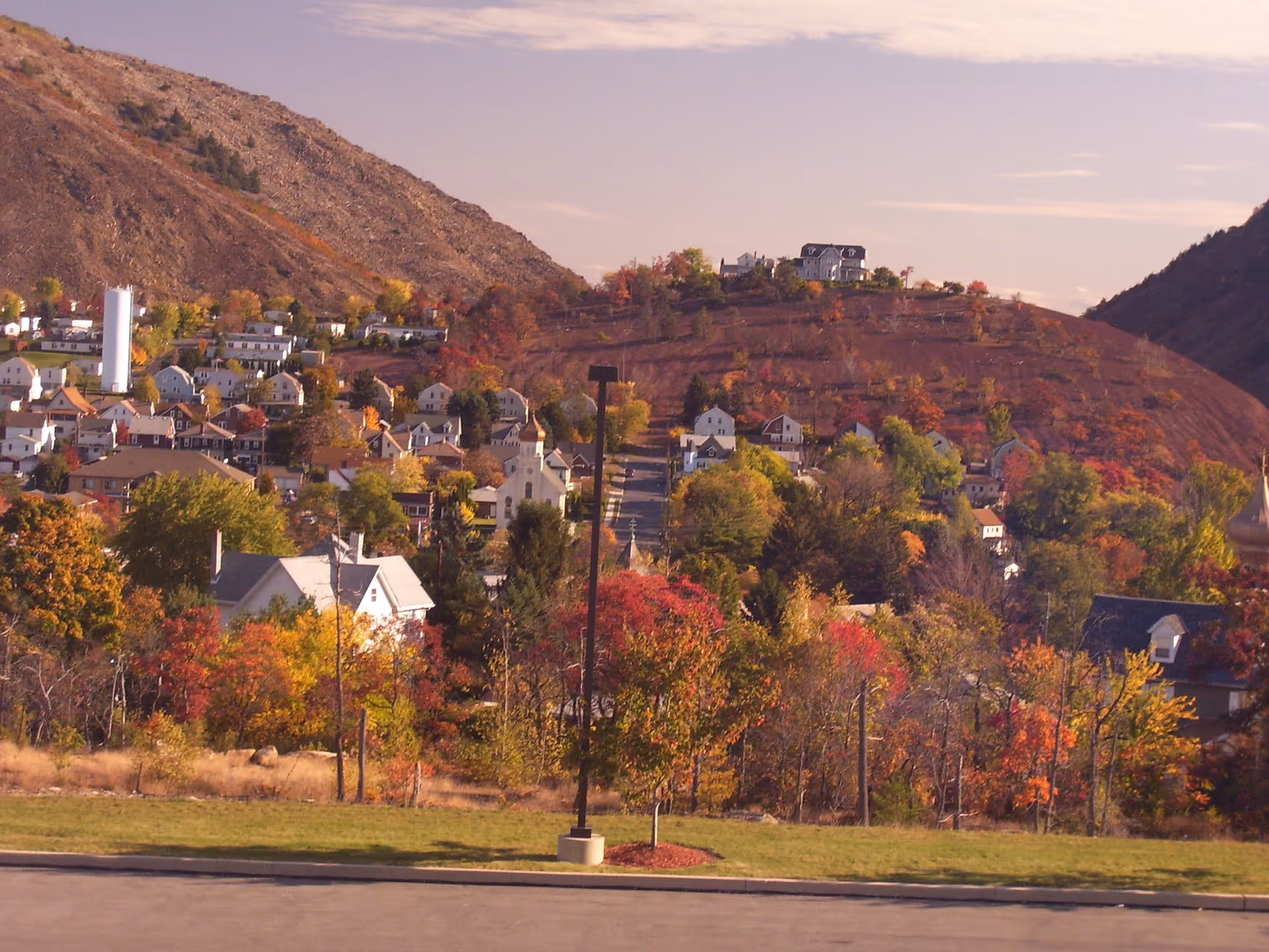 View of a small town nestled in a valley with houses scattered on the hillside surrounded by trees with autumn foliage. A large rocky hill rises in the background under a partly cloudy sky. In the foreground, there is a paved area with a single streetlamp and a grassy patch.