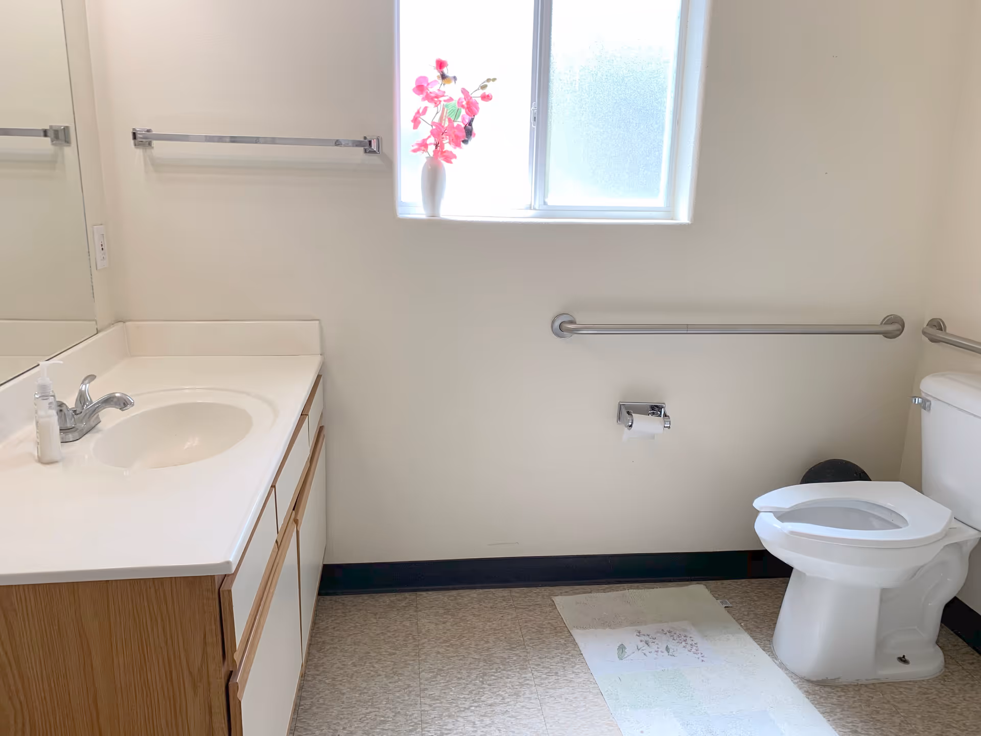 A clean bathroom with a white sink and countertop on the left, a large mirror above the sink, a window with frosted glass and a vase with pink flowers on the windowsill, a toilet with a raised seat on the right, grab bars on the walls, a toilet paper holder, and a small rug on the floor.