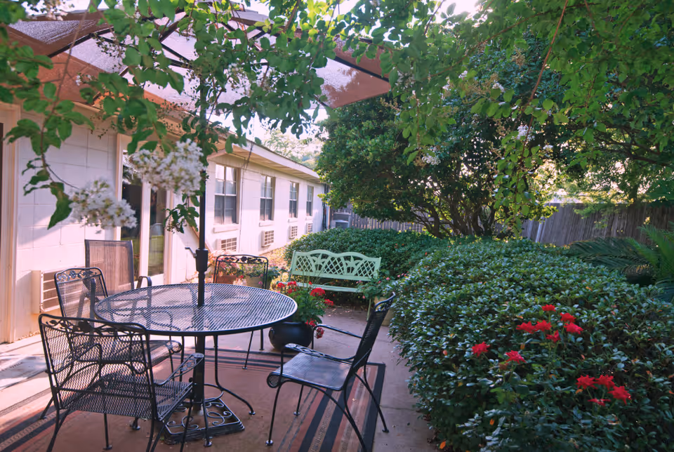 Outdoor patio area at Twin Oaks Nursing & Convalescent Home with a round metal table and four chairs under a large umbrella. Surrounding the patio are green bushes, trees, and red flowers, with a light green bench in the background near the building.