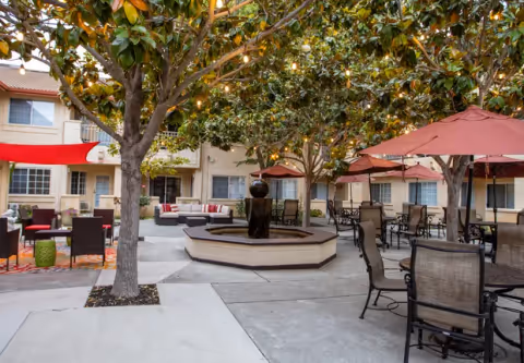 Outdoor courtyard with a central fountain, trees with string lights, patio seating and umbrellas in front of a multi-story building.