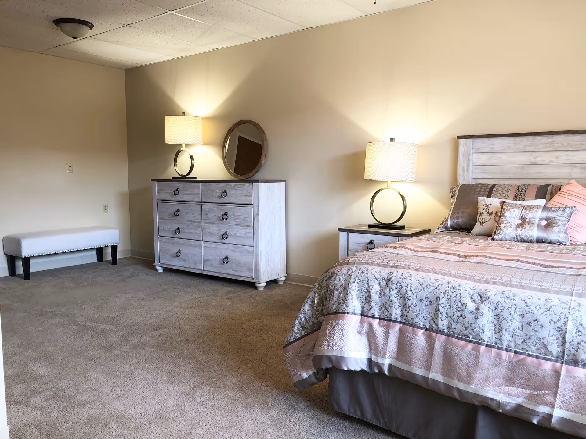 A cozy bedroom in a senior living facility featuring a bed with patterned bedding and multiple decorative pillows, a nightstand with a lamp, a dresser with a round mirror and another lamp, and a small upholstered bench against the wall. The room has beige walls and carpeted flooring.