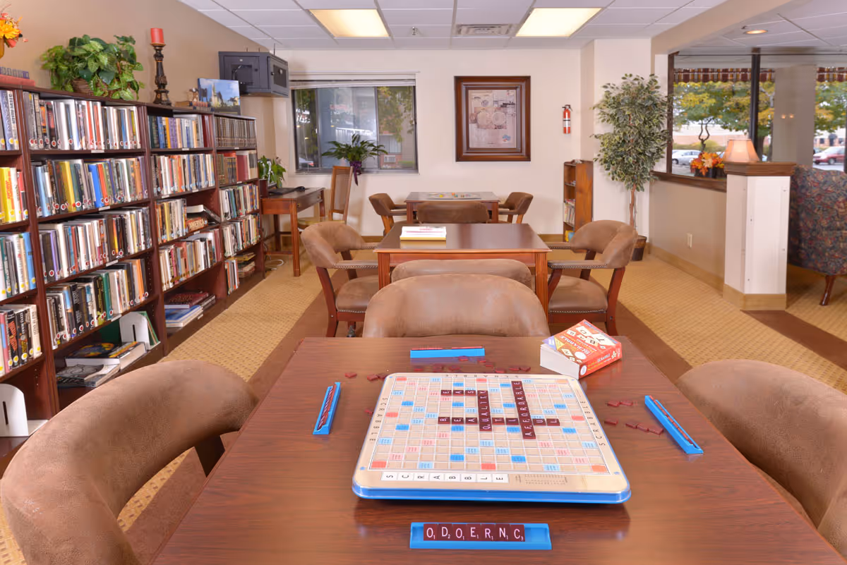 Cozy senior living common room with bookshelves, tables and a Scrabble game set up on the foreground table.
