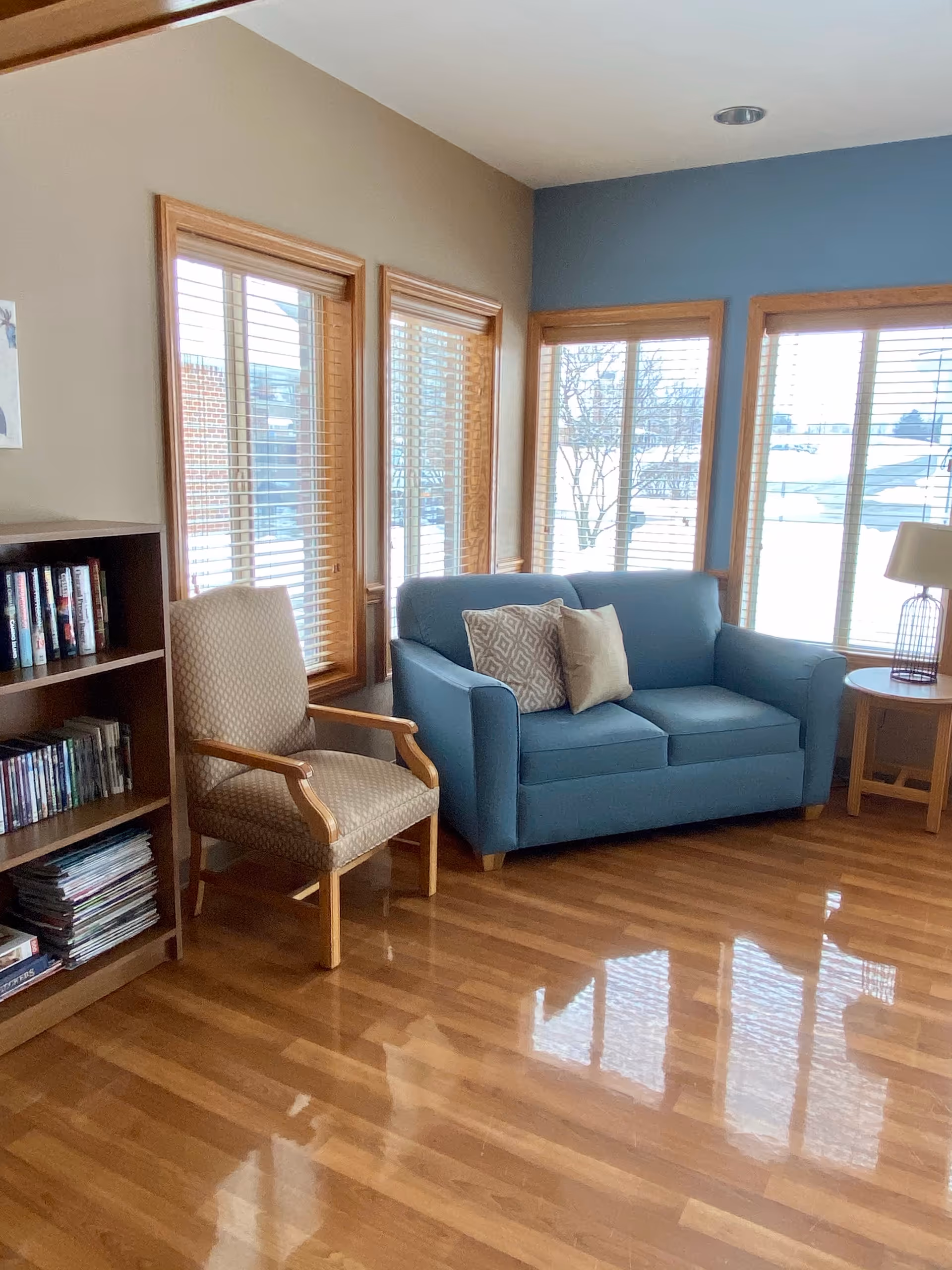A cozy living room area with a blue loveseat adorned with two decorative pillows, a beige armchair with wooden armrests, a wooden bookshelf filled with books and magazines, and large windows with wooden blinds letting in natural light. There is a small round wooden side table with a lamp on it next to the loveseat. The floor is polished wood.
