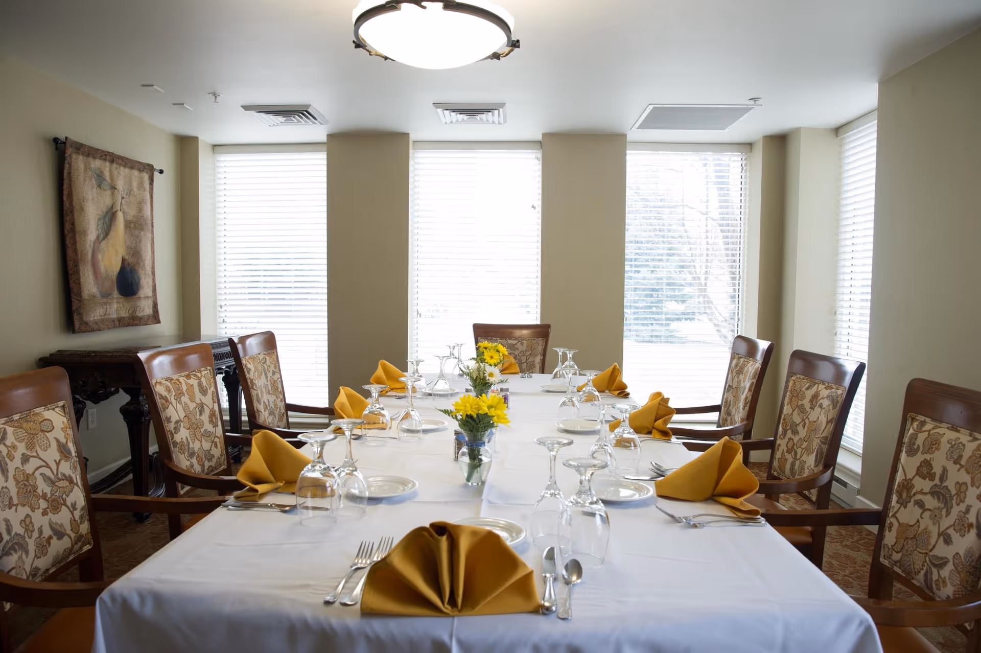 A formal dining room with a long rectangular table covered with a white tablecloth. The table is set with yellow folded napkins, glassware, plates, and silverware. There are eight upholstered wooden chairs around the table. The room has large windows with white blinds, a ceiling light fixture, and a decorative wall hanging on the left wall.