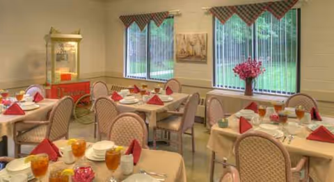 A dining room with several tables set for meals, pink chairs, red napkins, and windows.