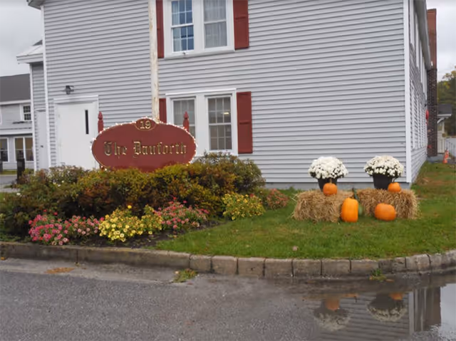 Front exterior of a white building with red shutters, a landscaped sign reading "The Danforth", and autumn decorations of pumpkins, hay bales, and potted flowers.