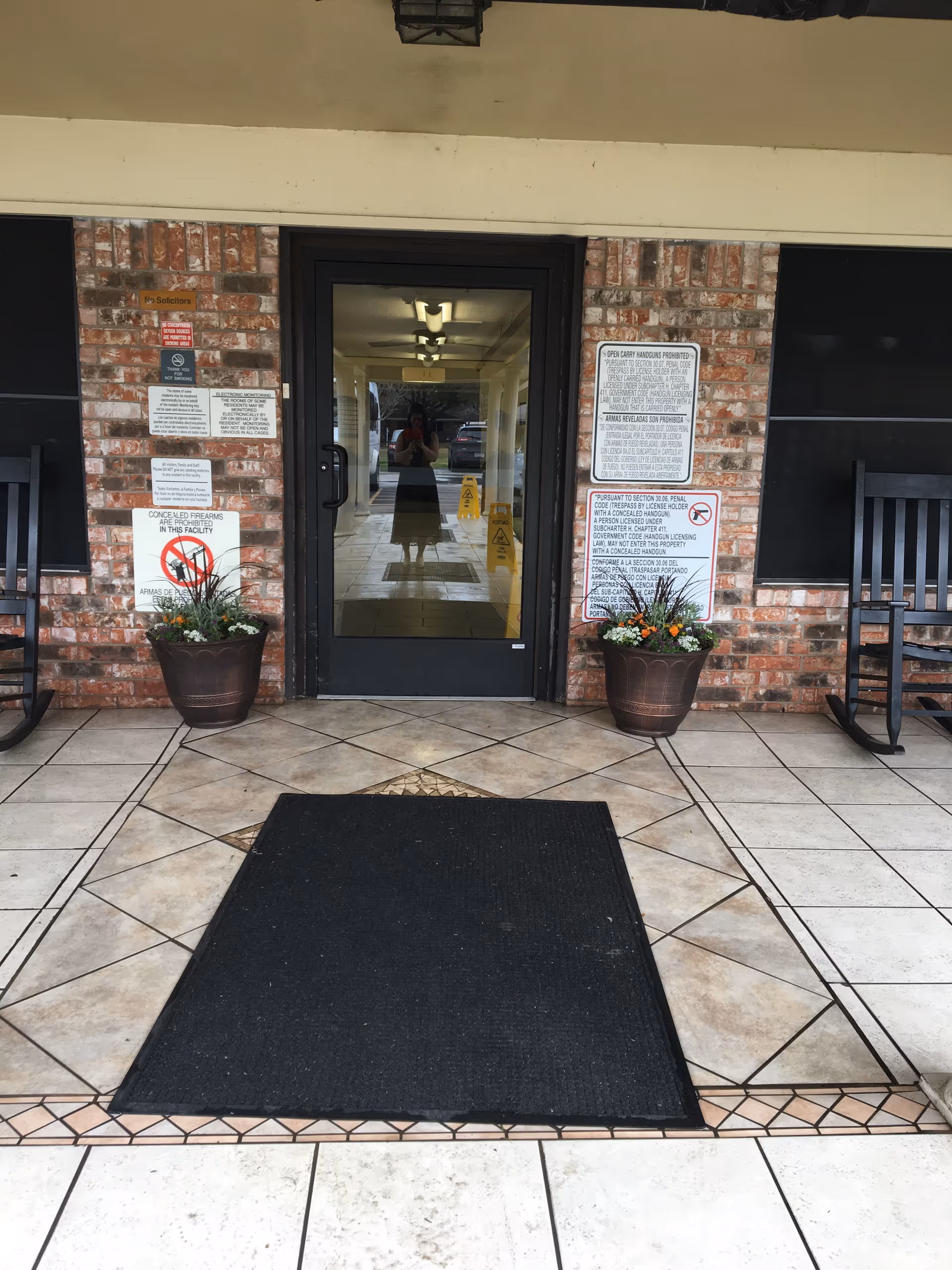 Entrance to a building with a glass door framed by brick walls. There are two potted plants with flowers on either side of the door, two black rocking chairs, and several signs posted on the brick walls. A black mat is placed on the tiled floor in front of the door.