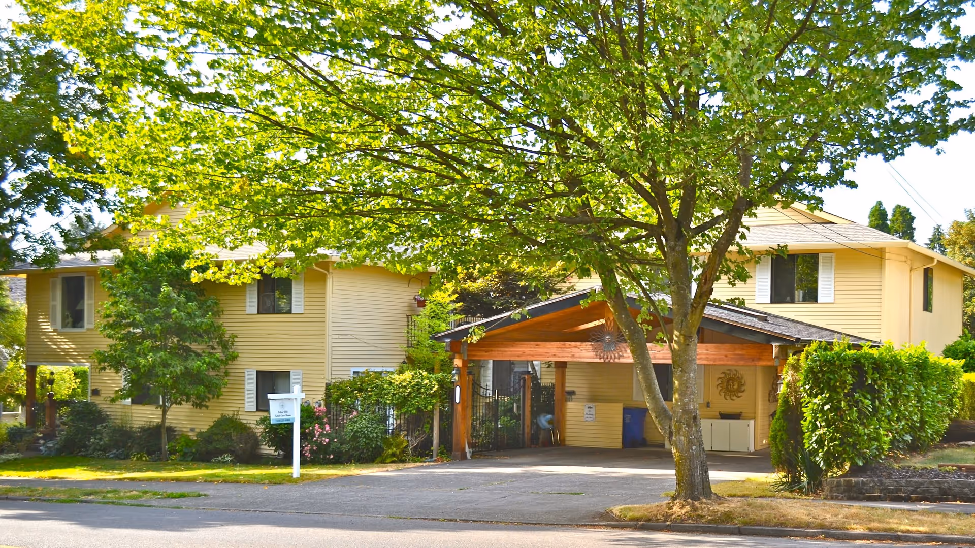 A two-story yellow building with white window shutters partially obscured by green trees and bushes. The building has a covered driveway with wooden beams and a gated entrance. There is a white signpost on the lawn in front of the building.