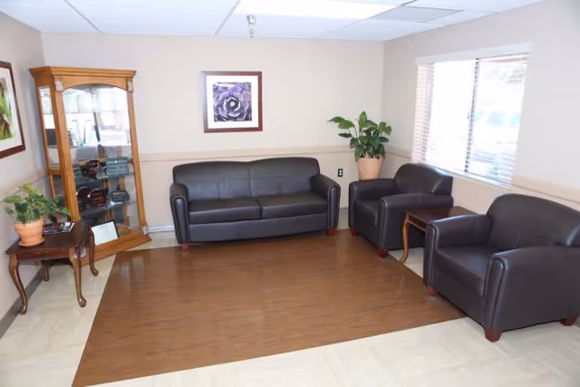 A small waiting room with a black leather sofa and two matching armchairs, a wooden display cabinet, potted plants, and a window.