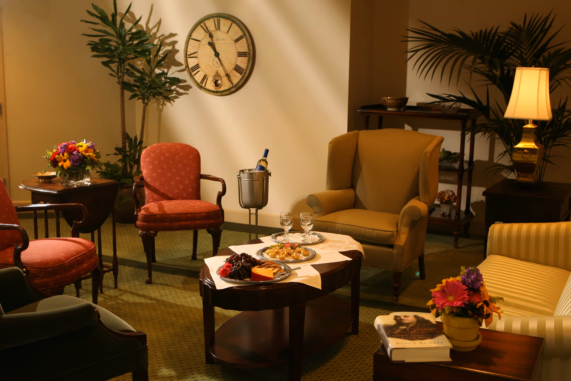 A cozy living room with a green armchair, two red upholstered chairs, and a striped sofa. A wooden coffee table in the center holds plates of snacks, two glasses, and a bottle of wine in an ice bucket. There are plants, a wall clock, a side table with a lamp, and a small table with a flower vase and a book.