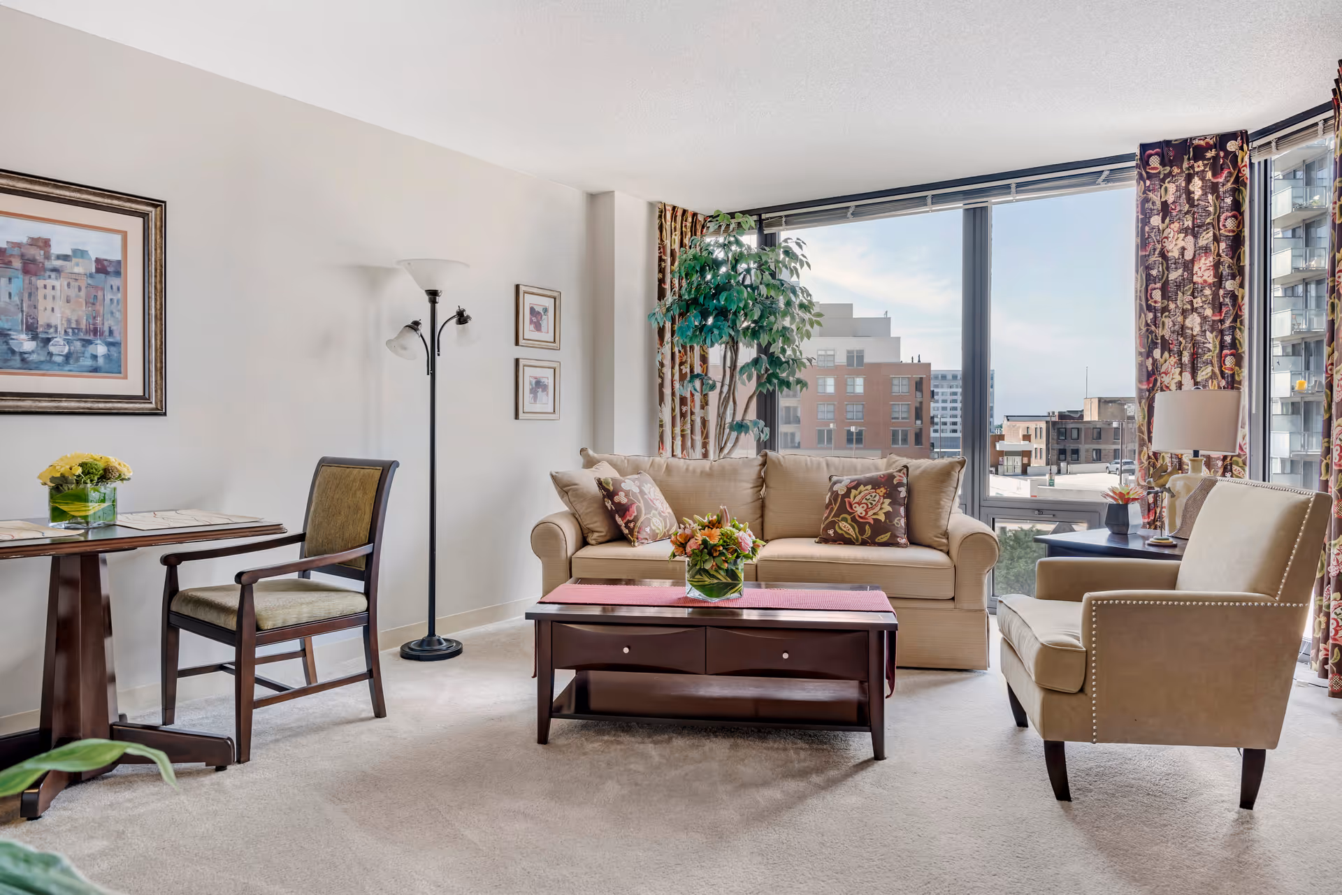 A bright living room with a beige sofa adorned with patterned pillows, a matching armchair, a dark wooden coffee table with a flower arrangement, a small dining table with a chair, a floor lamp, and large windows with floral curtains showing a cityscape outside.
