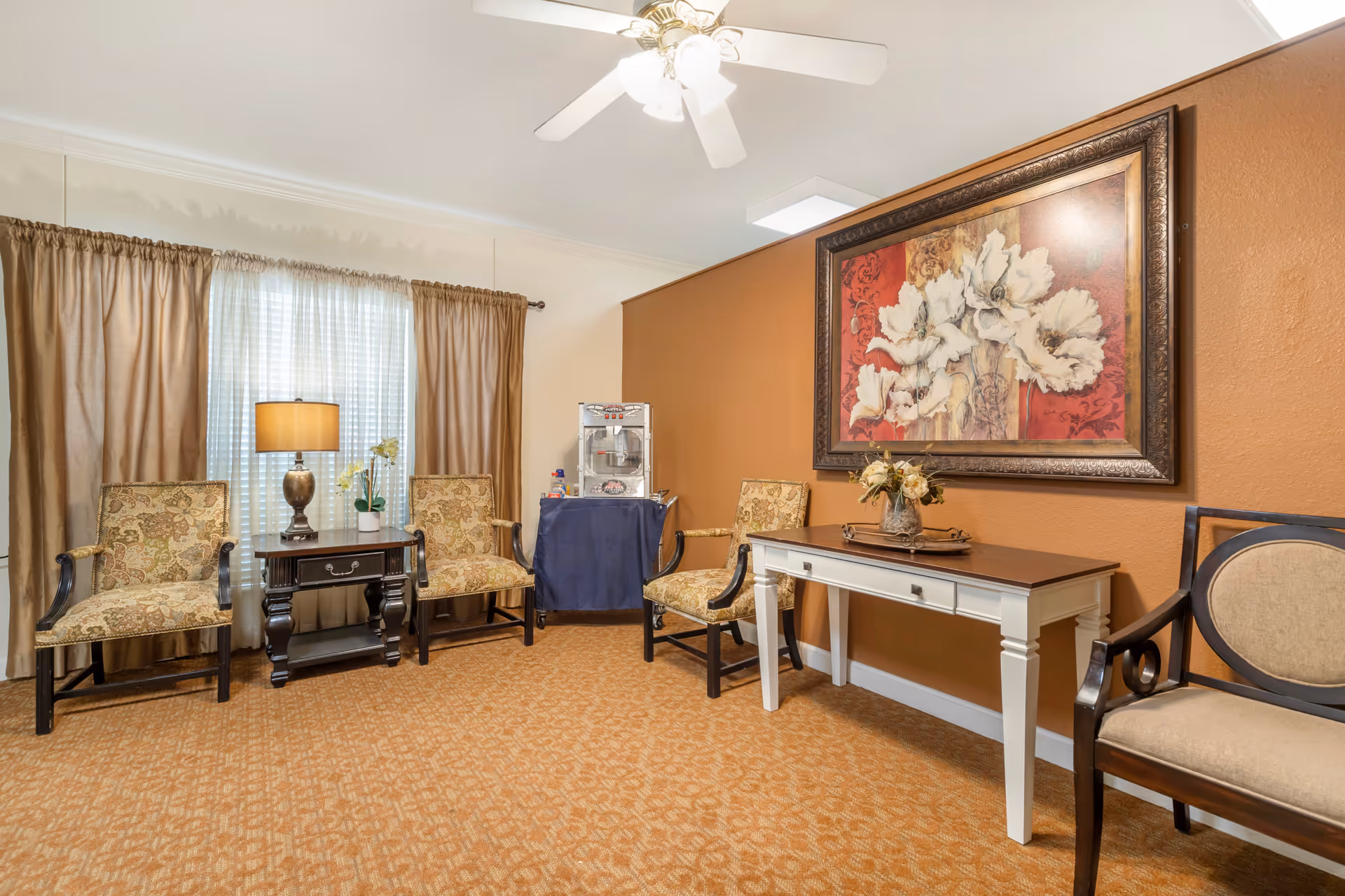A cozy sitting area with four upholstered chairs arranged around a small dark wooden side table with a lamp and a potted orchid. There is a white console table with a floral arrangement on top against a burnt orange wall, which also features a large framed painting of white flowers. A popcorn machine is placed on a small table covered with a blue cloth in the corner. The room has beige curtains and a ceiling fan with lights.