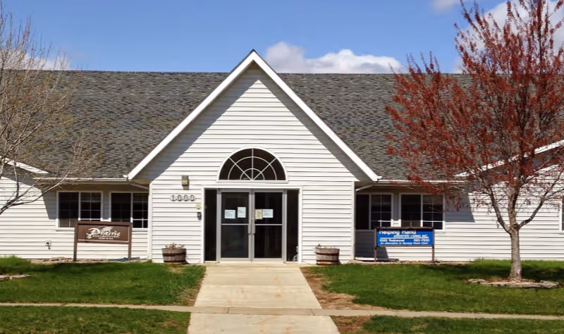 White single-story building front with a gabled entrance, double glass doors, windows, and trees on a grassy lawn.