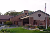 Exterior view of a single-story brick building with a covered entrance, landscaped garden with flowers and shrubs, and an American flag on a flagpole in front of the building under a partly cloudy sky.