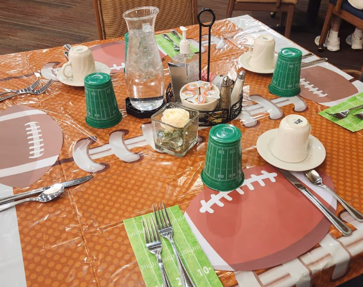 Dining table set with football-themed tablecloth and placemats, cups, utensils, a water carafe, and a small centerpiece.