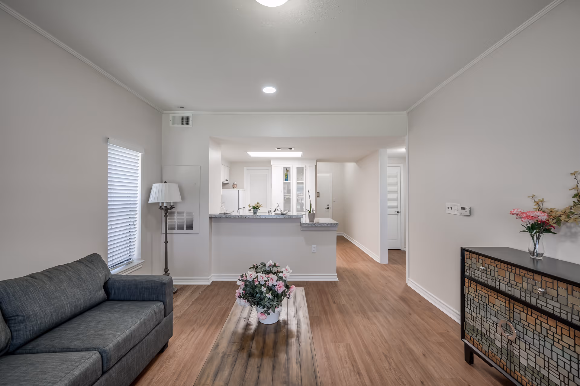 A modern living room with a gray sofa on the left, a wooden coffee table with a flower pot in the center, and a decorative cabinet with flowers on the right. The room has light-colored walls, wood flooring, and a window with blinds. The living room opens into a kitchen area with white cabinets and a countertop.