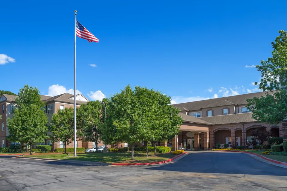 Exterior view of Town Village Tulsa senior living facility with a large American flag on a flagpole, surrounded by green trees and a clear blue sky. The building is made of brick with multiple windows and an entrance driveway.