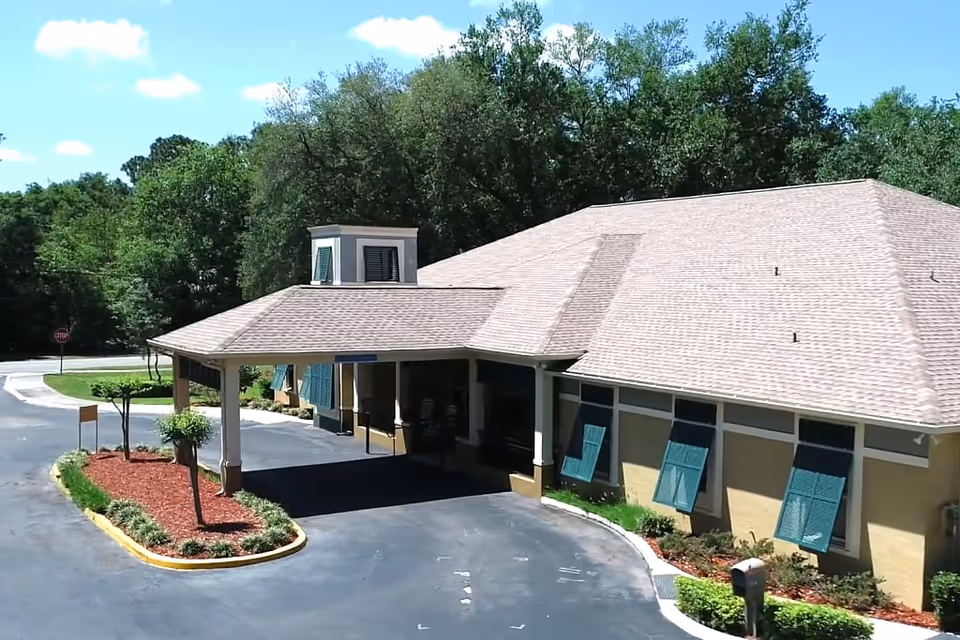Exterior view of a single-story building with a covered entrance driveway, surrounded by landscaped greenery and trees under a blue sky with some clouds.