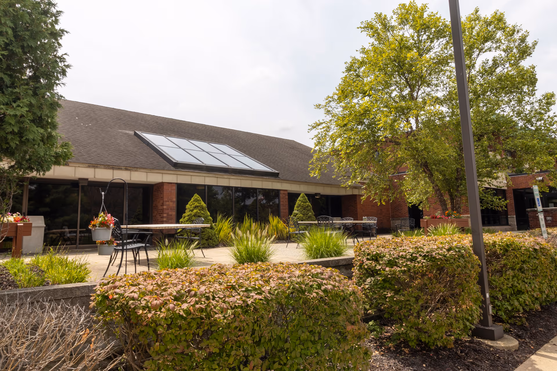 Outdoor patio area at StoryPoint Grand Rapids East with tables and chairs surrounded by greenery, bushes, and trees in front of a brick building with large windows and a sloped roof with skylights.