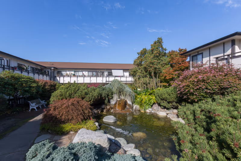 A landscaped outdoor garden area at a senior living facility featuring a small pond with rocks and a waterfall, surrounded by various shrubs, trees, and flowering plants. The building with balconies and windows is visible in the background under a clear blue sky.