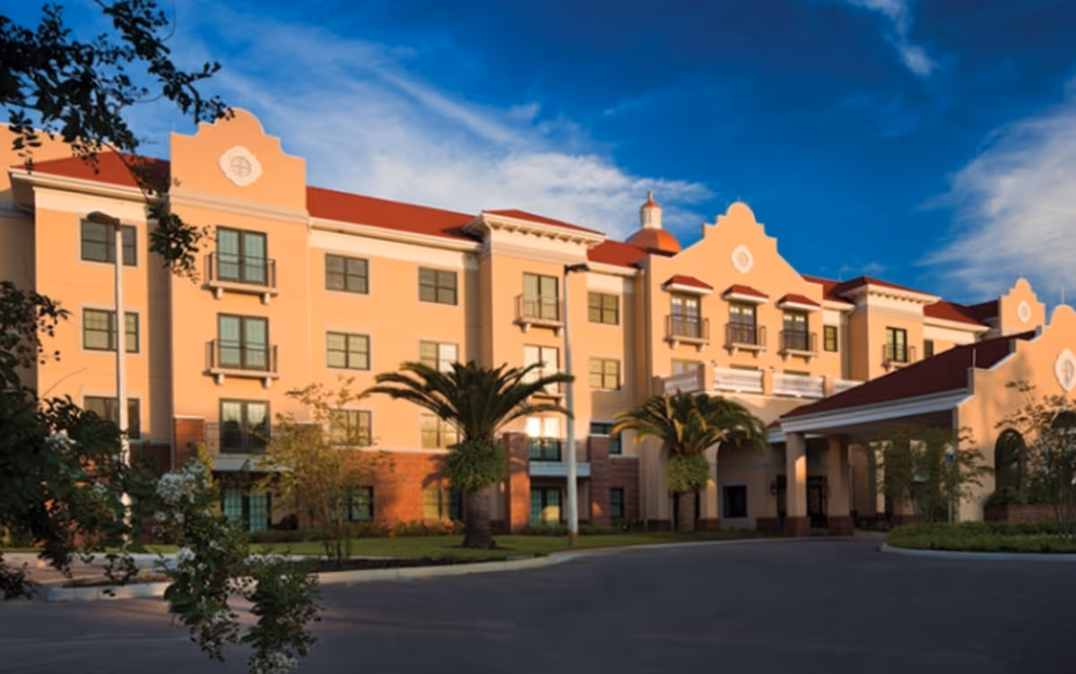 Front exterior of a Mediterranean-style four-story senior living building with palm trees and a covered entrance under a blue sky.
