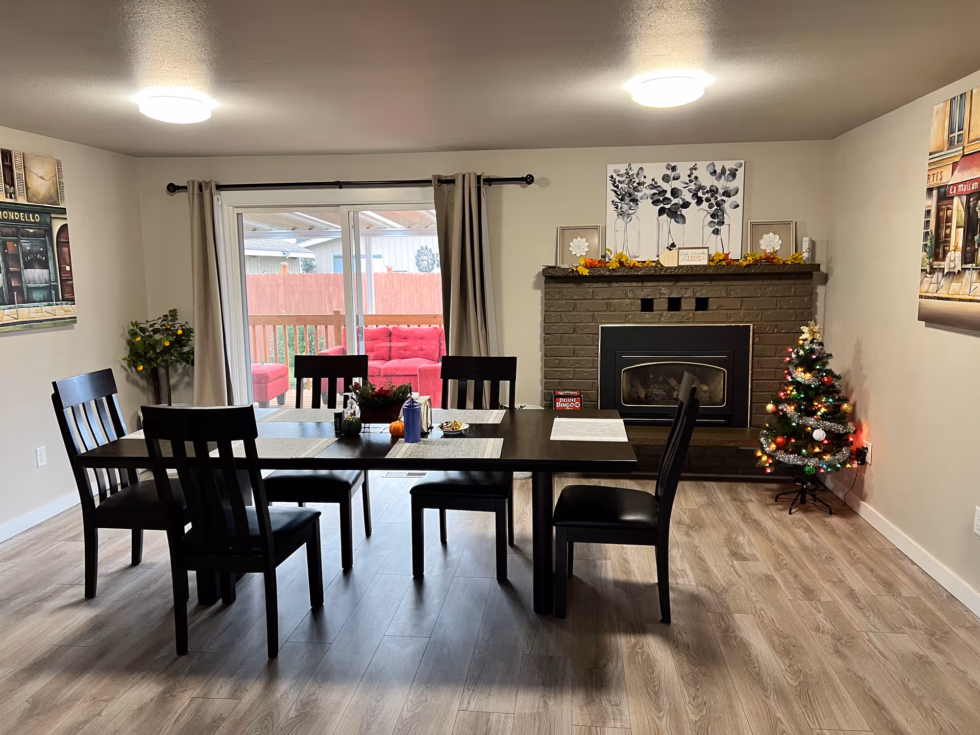 Dining room with a dark wood table and six chairs, a fireplace and small decorated Christmas tree, sliding glass door to a patio with red seating, and wall art.
