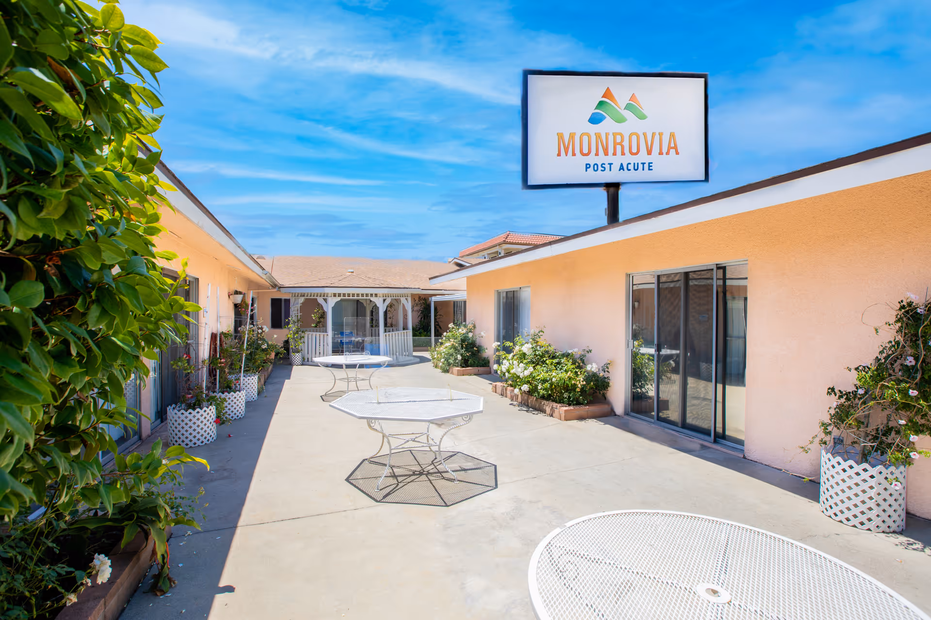 Outdoor courtyard area of Monrovia Post Acute facility with several white metal tables, potted plants along the walls, and a gazebo in the background under a blue sky.