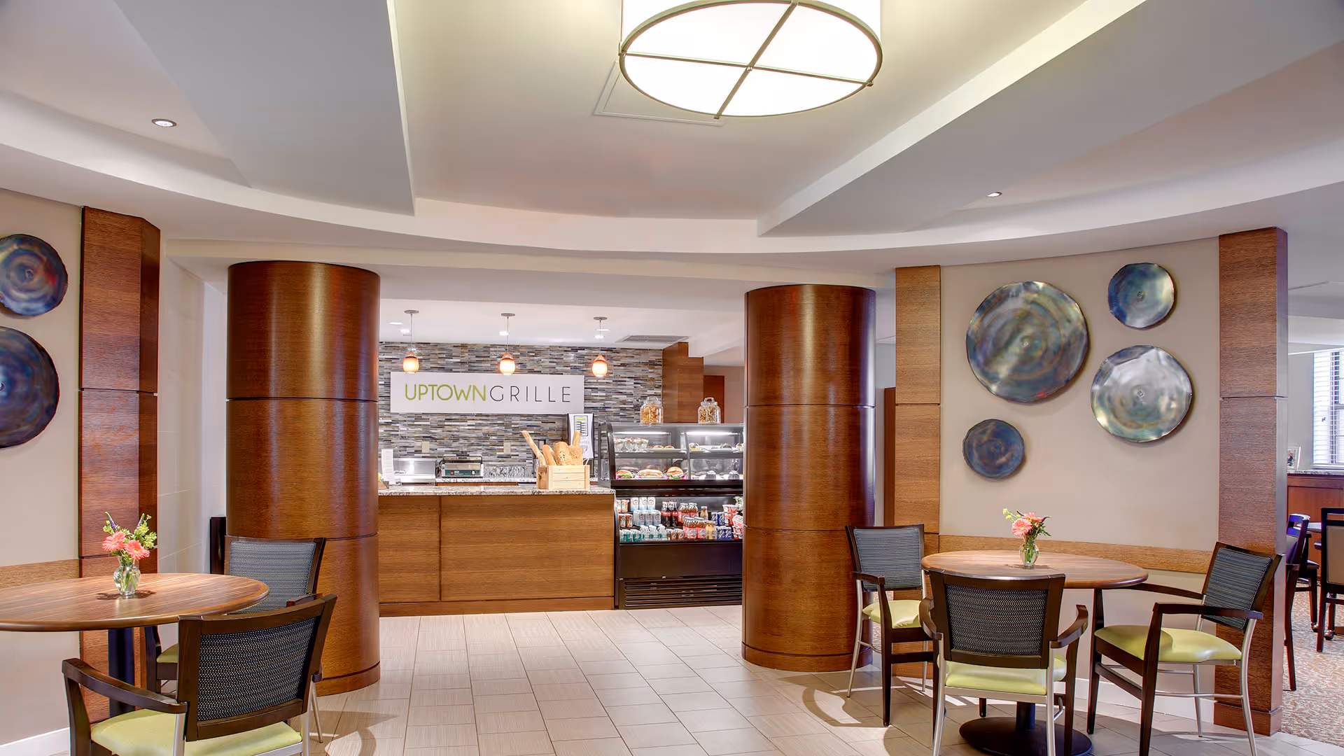 Interior view of a dining area named Uptown Grille with round wooden tables and chairs with green cushions. The space features two large wooden columns, decorative wall plates, and a counter displaying food and beverages under warm lighting.
