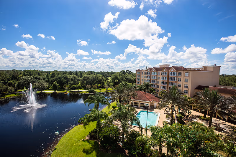 A multi-story residential building beside a lake with a fountain, a swimming pool, palm trees, and a bright blue sky.