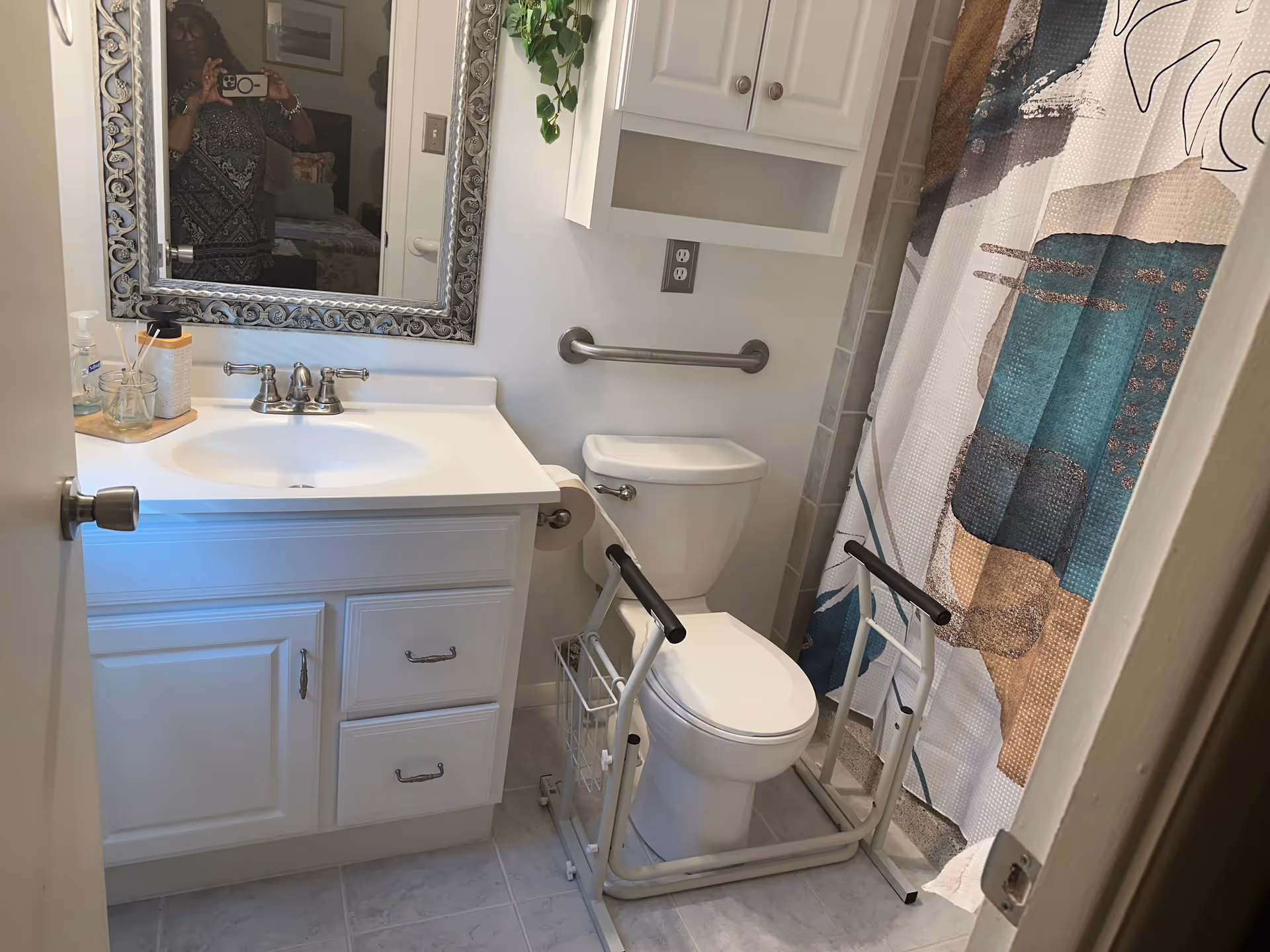 A small bathroom with a white vanity and sink, a decorative framed mirror above the sink, a toilet with a safety frame around it, a grab bar on the wall, and a shower with a colorful patterned curtain. A person is partially visible in the mirror taking the photo.