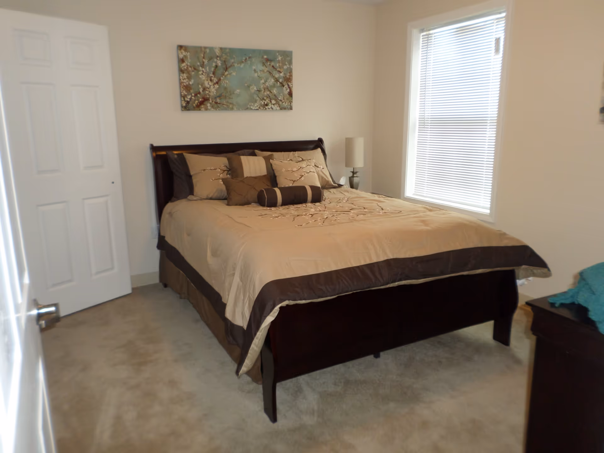 A bedroom with a neatly made bed featuring beige and brown bedding and multiple pillows. There is a window with blinds on the right wall, a nightstand with a lamp next to the bed, and a floral painting hanging on the wall above the headboard. The room has beige carpet and light-colored walls.