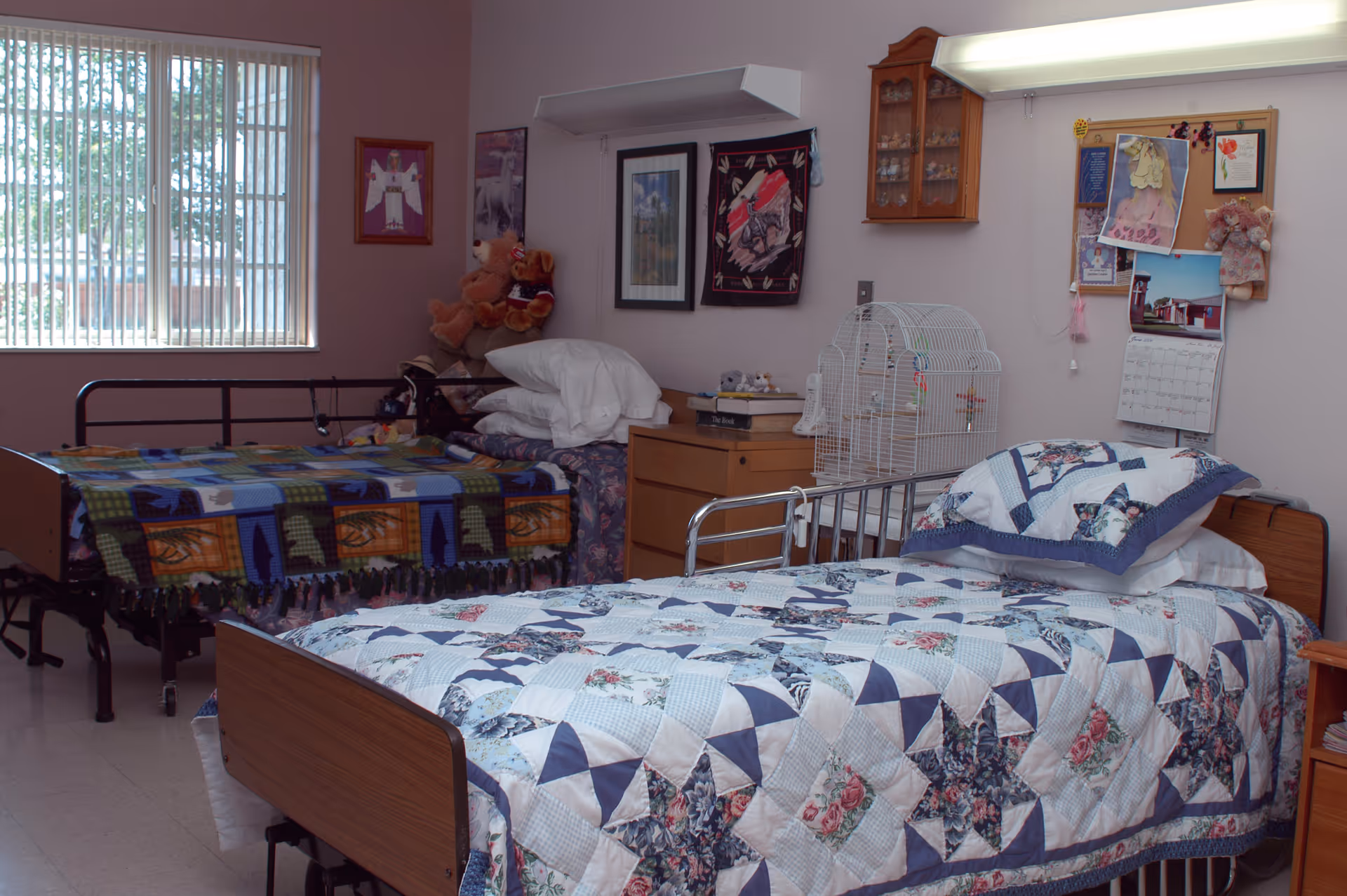 A room with two hospital-style beds, each covered with colorful quilts. One bed has a blue and white star-patterned quilt, and the other has a multicolored quilt with animal designs. There is a window with vertical blinds on the left wall, a wooden dresser between the beds with a birdcage on top, and various decorations including framed pictures, a calendar, and stuffed animals on the walls.