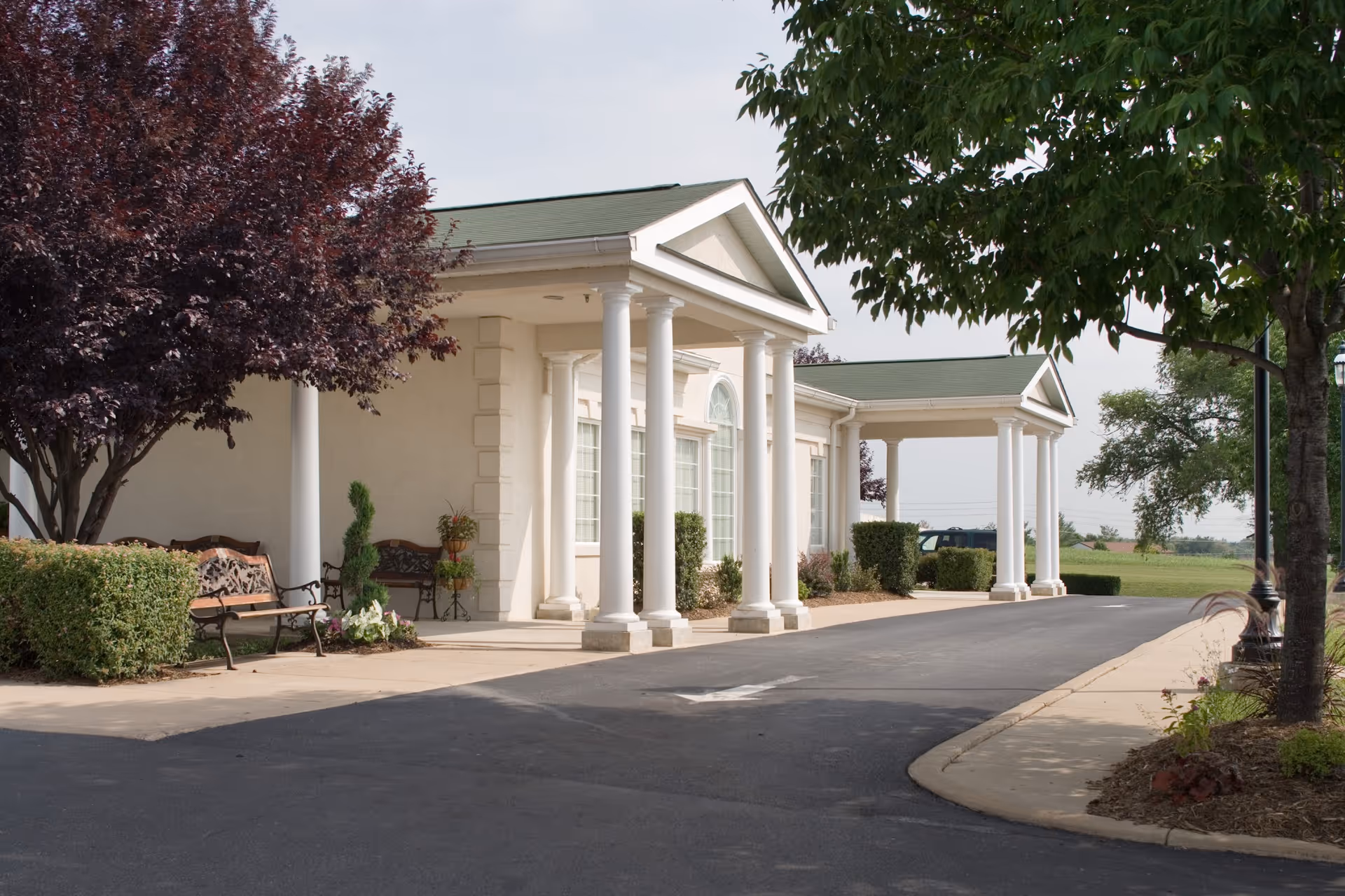 Front entrance of a single-story building with white columns, a covered portico, benches, a driveway, and surrounding trees and shrubs.